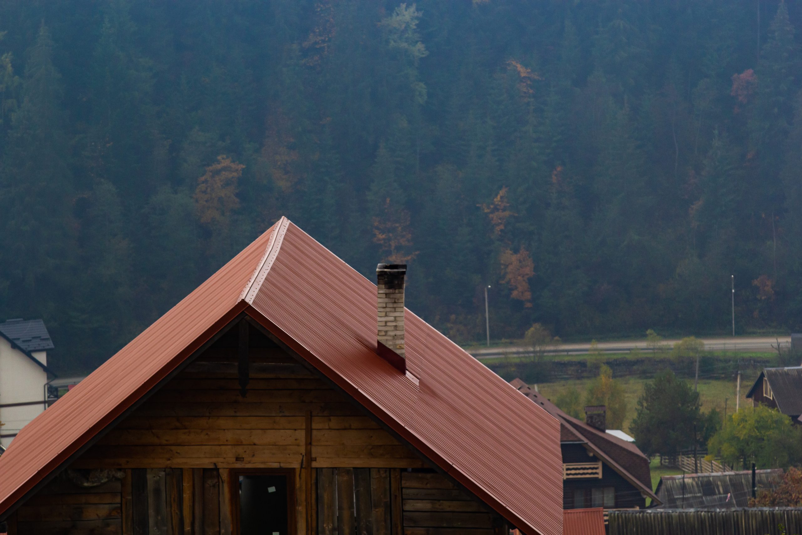 House With New Brown Metal Tile Roof And Rain Gutter. Metallic G Red metal tile roof with newly installed gutter drainage system with the forrest in the background